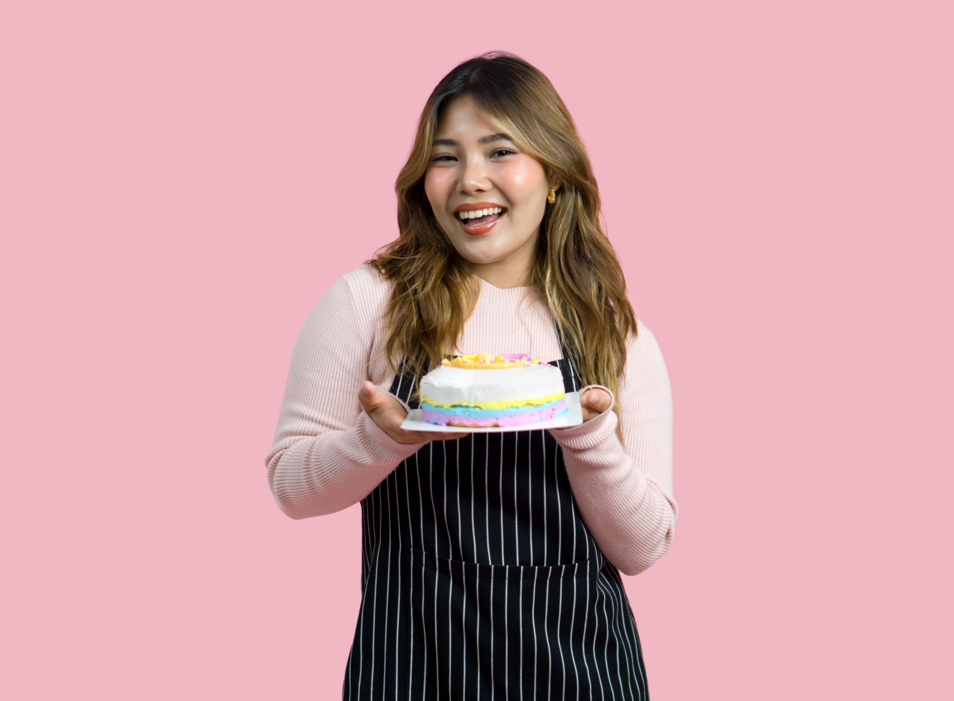 Young asian woman with an apron in celebratory or festive mood holding colorful cake with various sprinkles on the topping. Portrait on pink background with studio light. Isolated