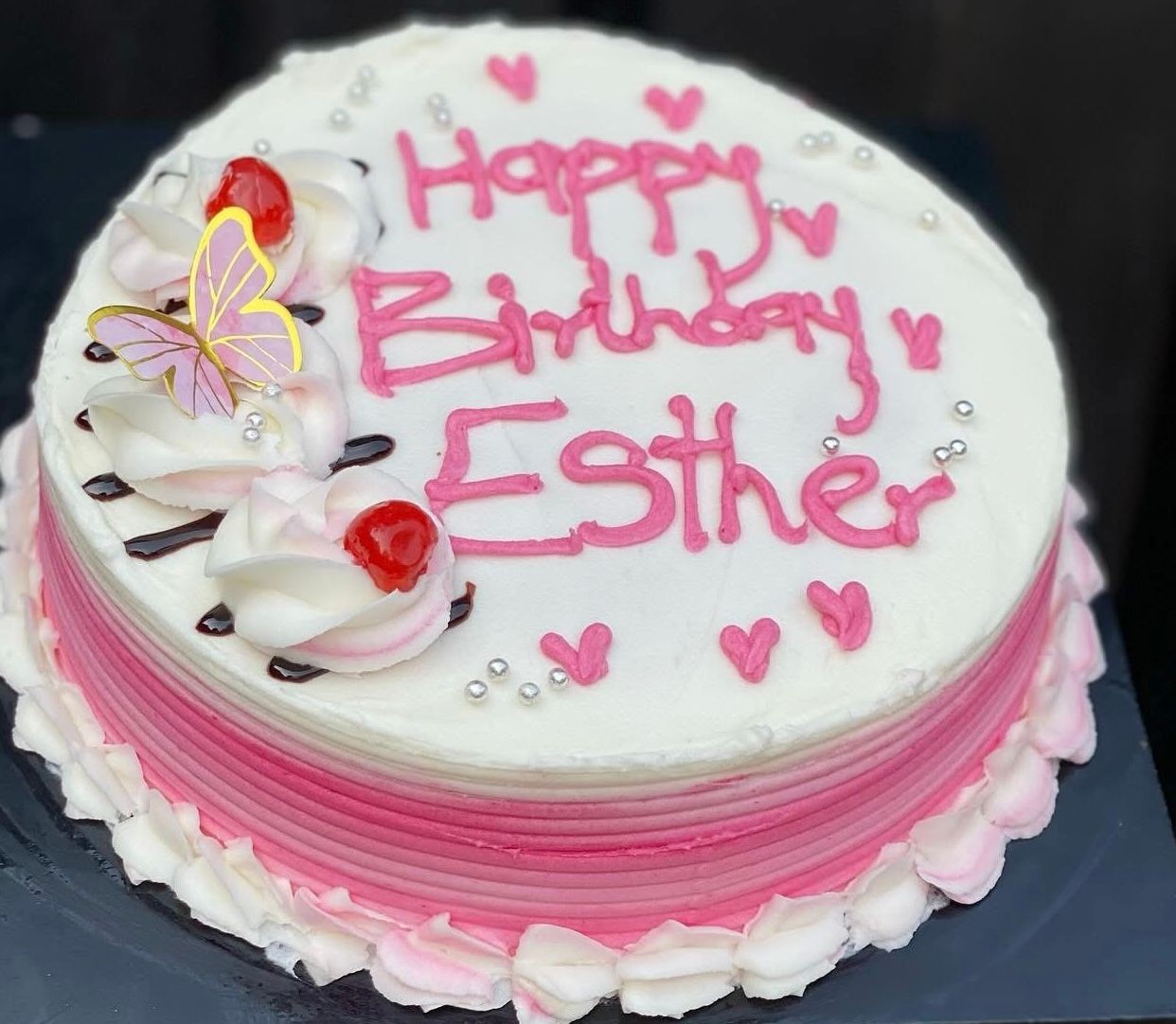 Birthday cake with pink icing reading 'Happy Birthday Esther,' decorated with cherries and a butterfly topper.