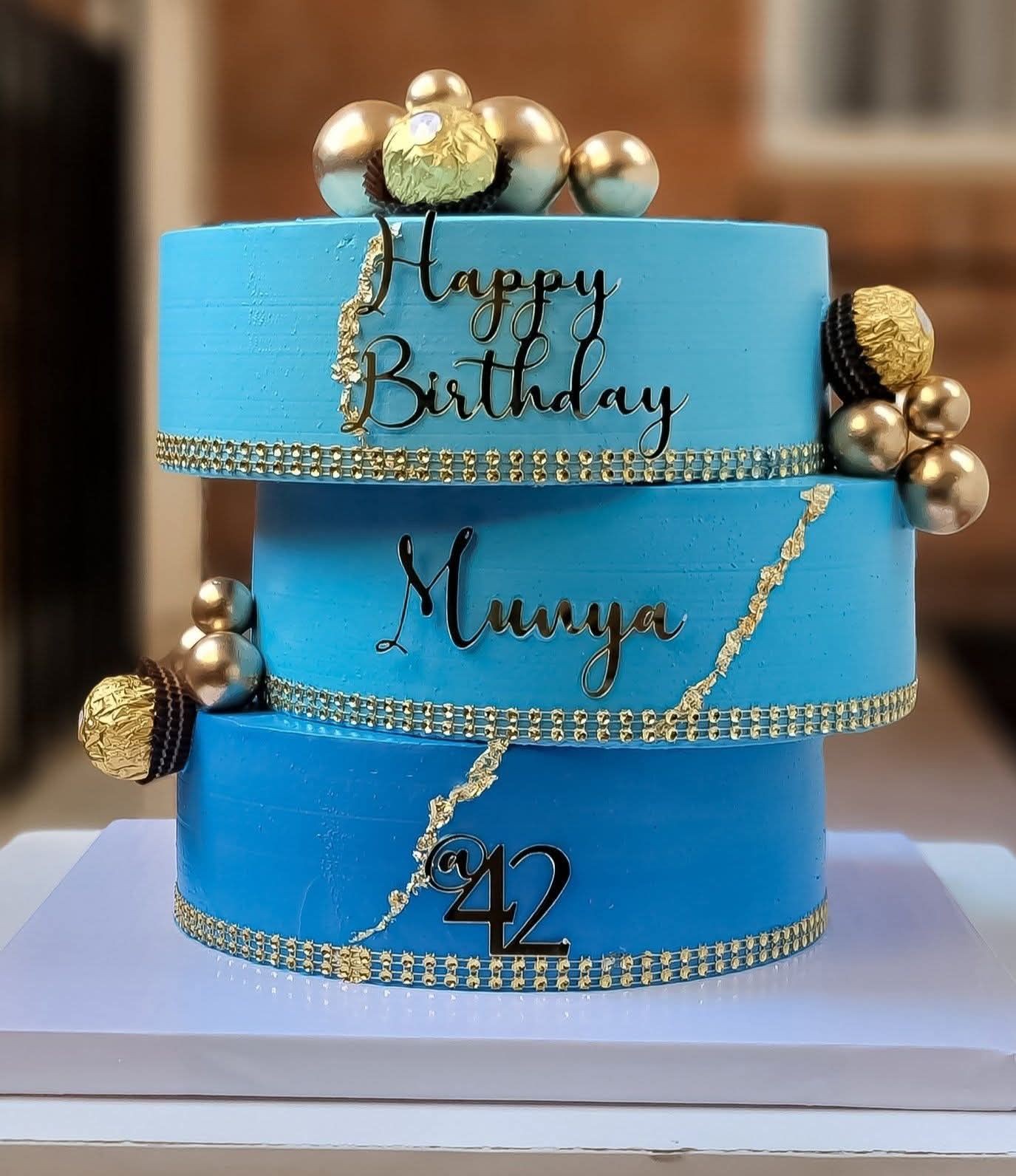 Blue two-tier cake with chocolates and gold decorations, text reads Happy Birthday Munya @42.