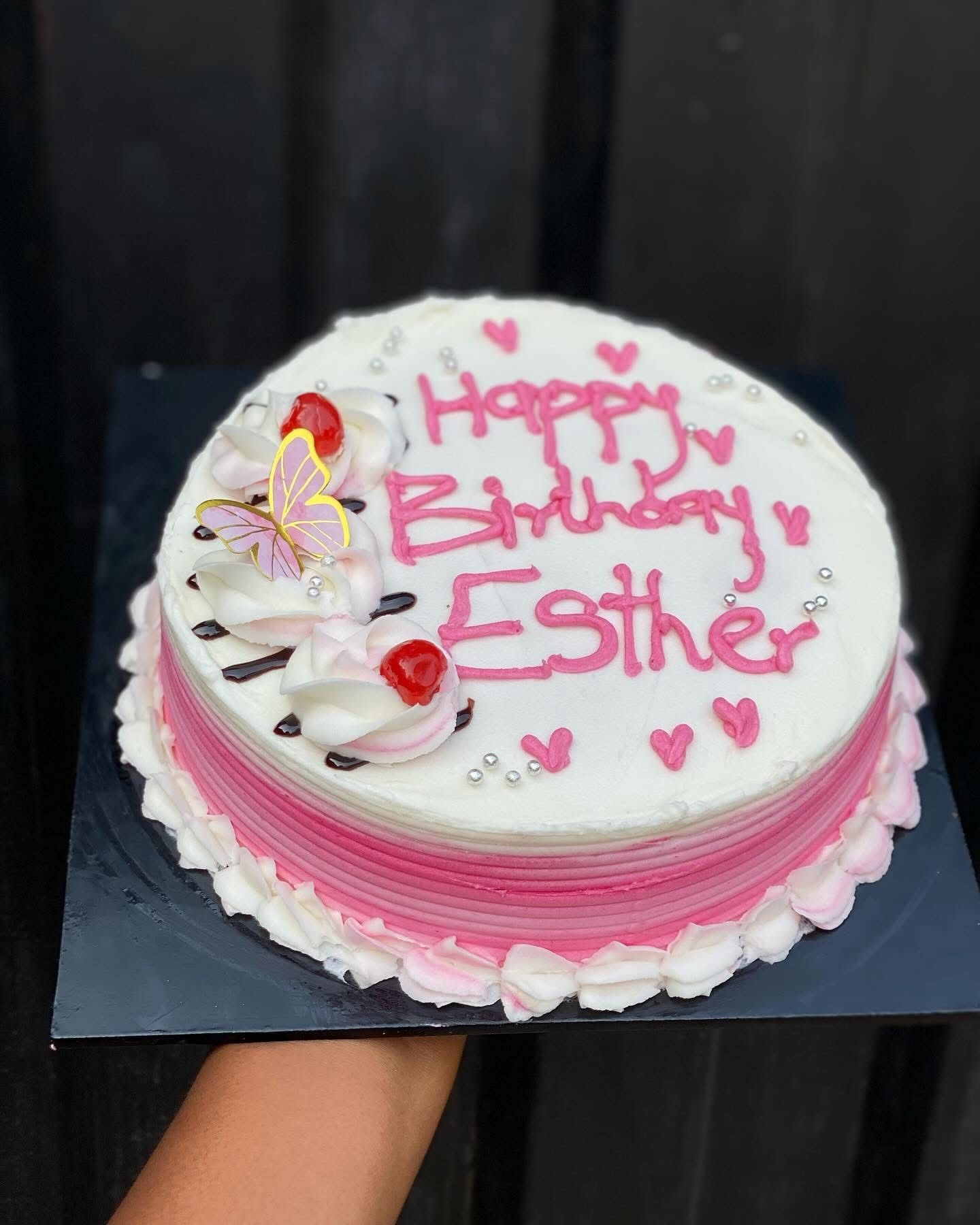 Round pink and white birthday cake with cherries and butterfly decorations on a dark background.