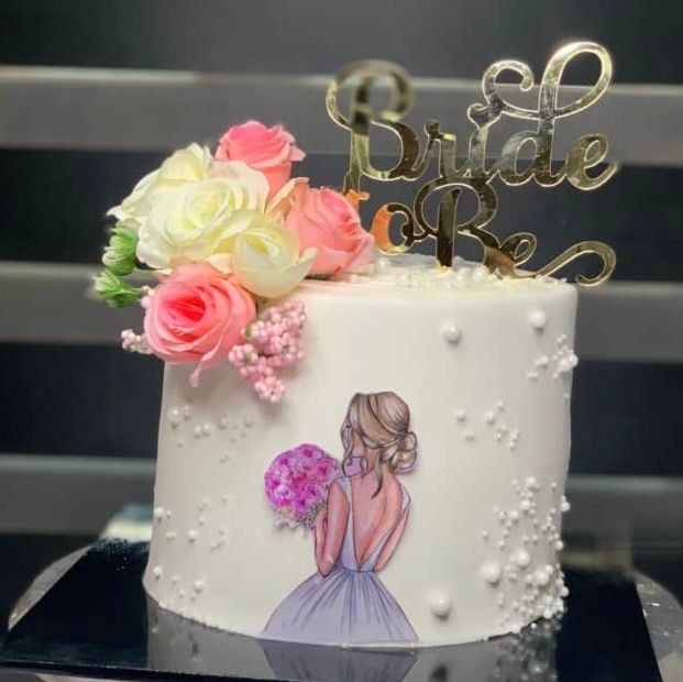 Elegant white cake with 'Bride to Be' topper, adorned with flowers and a bride illustration.