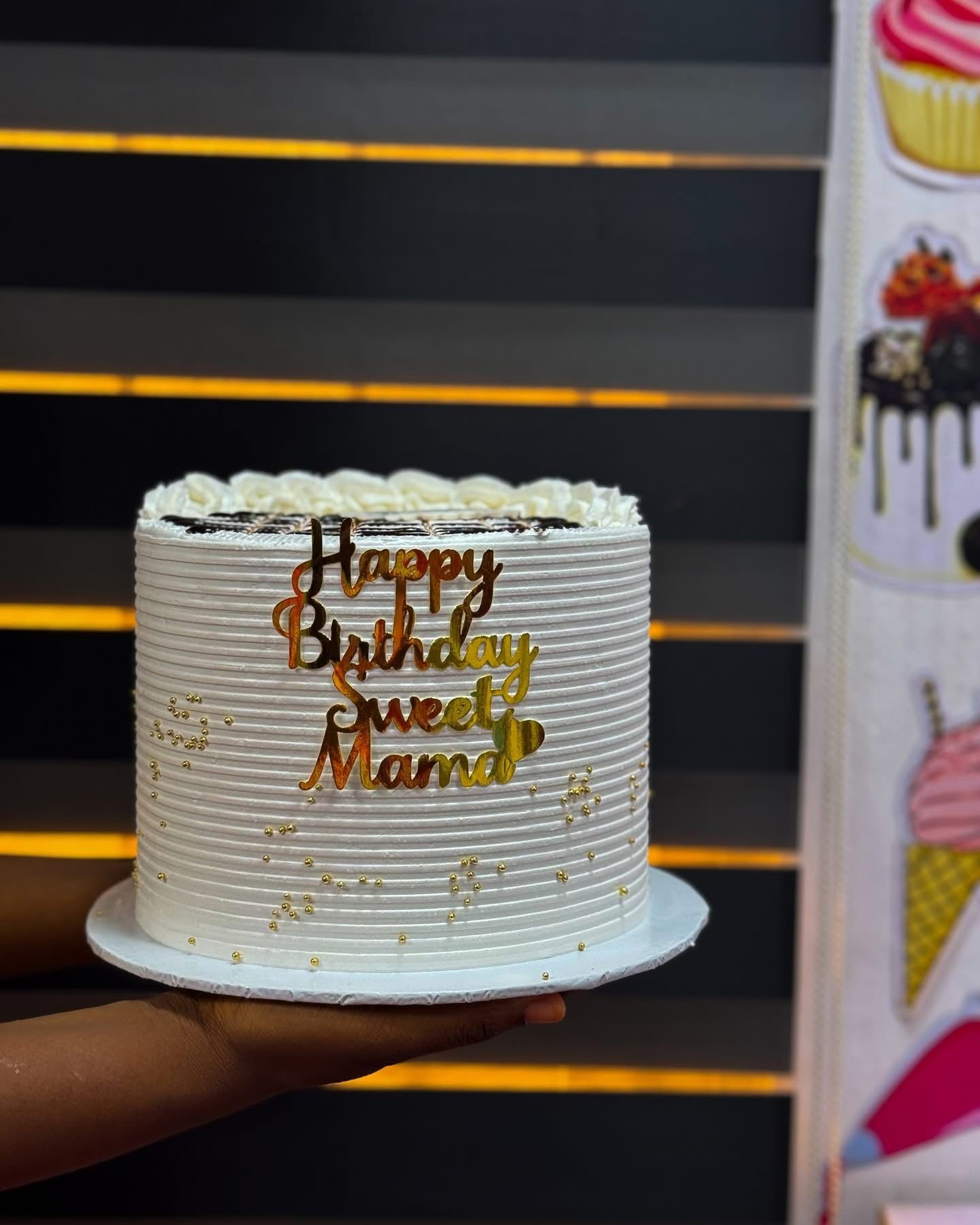 White birthday cake with gold text on a dark striped background, decorated with small gold dots.