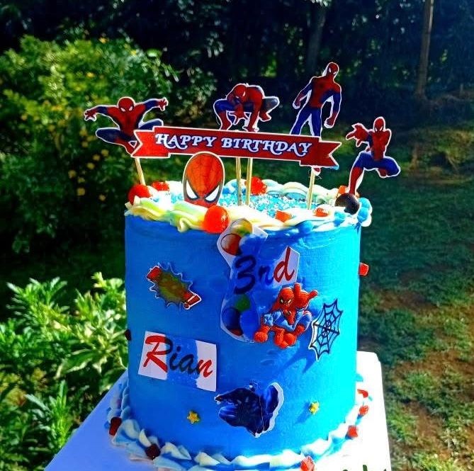 Blue superhero-themed birthday cake with figures and decorations celebrating a 3rd birthday outdoors.