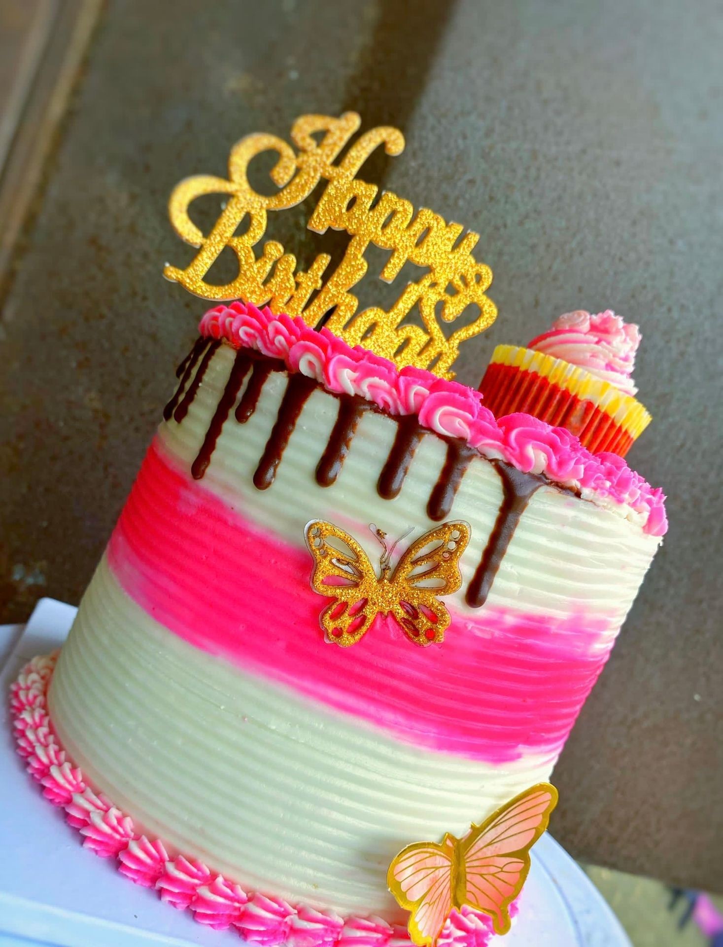 Pink and white birthday cake with chocolate drizzle, butterfly decorations, and 'Happy Birthday' topper.