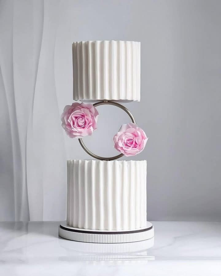 Tiered white cake with fluted design, decorated with pink roses on a metallic ring centerpiece.