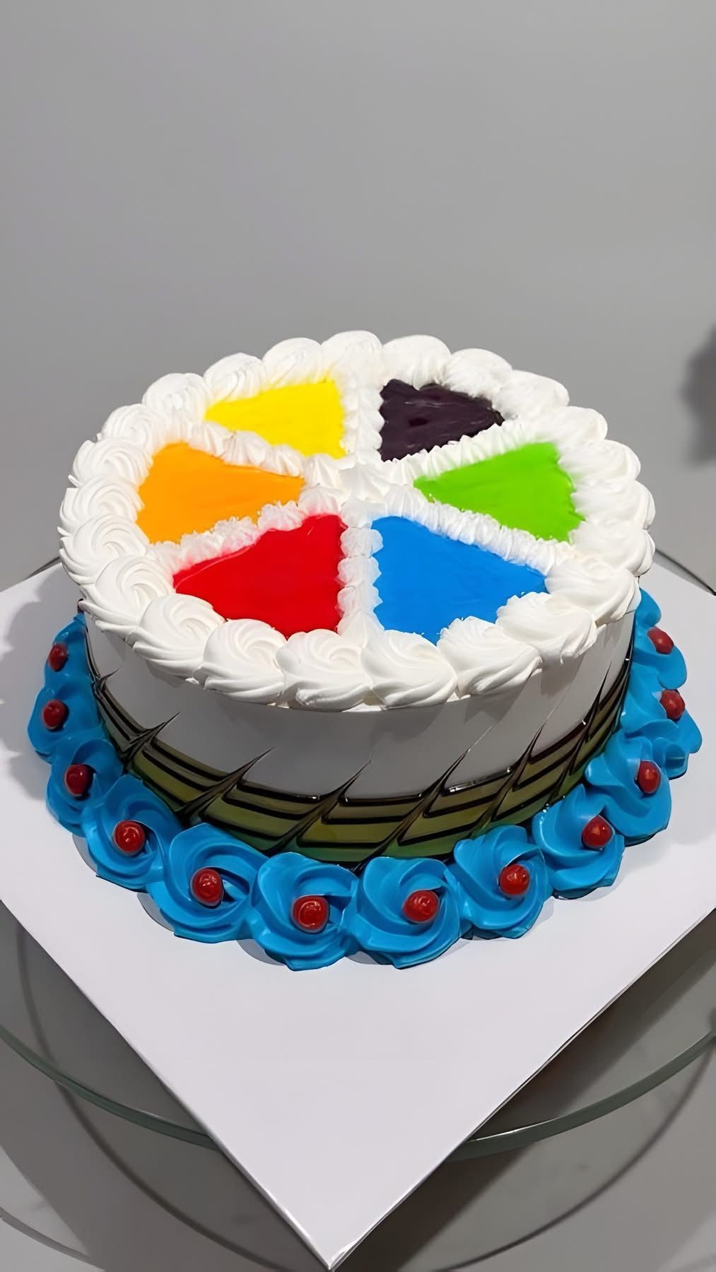 Colorful cream cake with rainbow segments and blue floral icing at the base on a glass table.