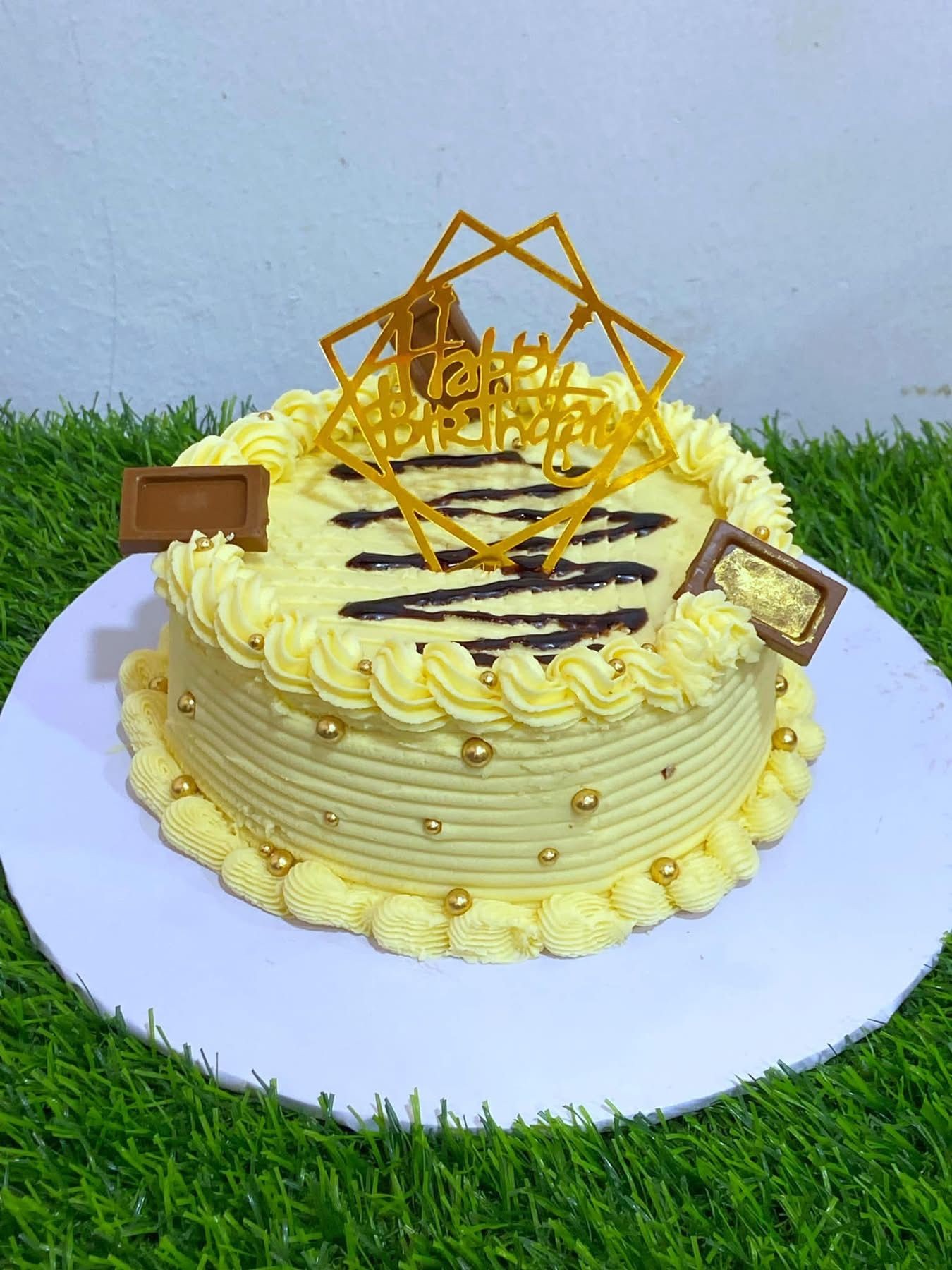 Yellow birthday cake with gold decorations and a topper on grassy surface.