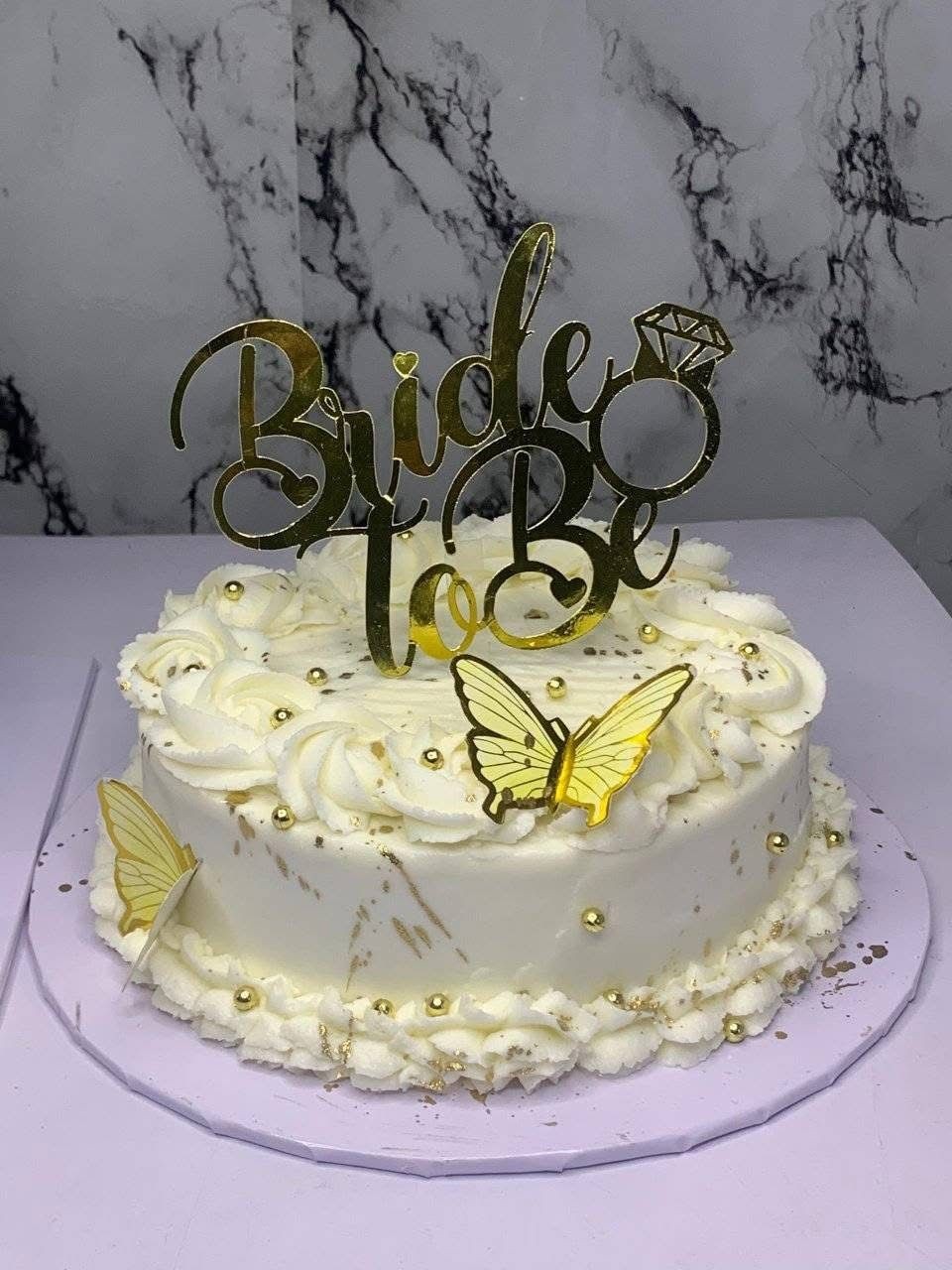 White cake with 'Bride to Be' topper and butterfly decorations on a marble backdrop.