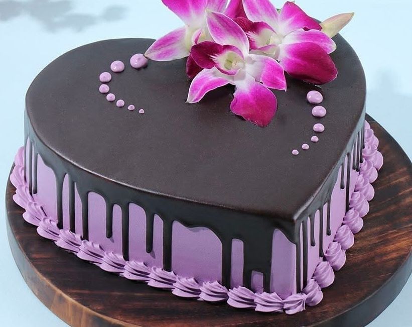 Heart-shaped cake with chocolate glaze and purple frosting, topped with pink flowers.