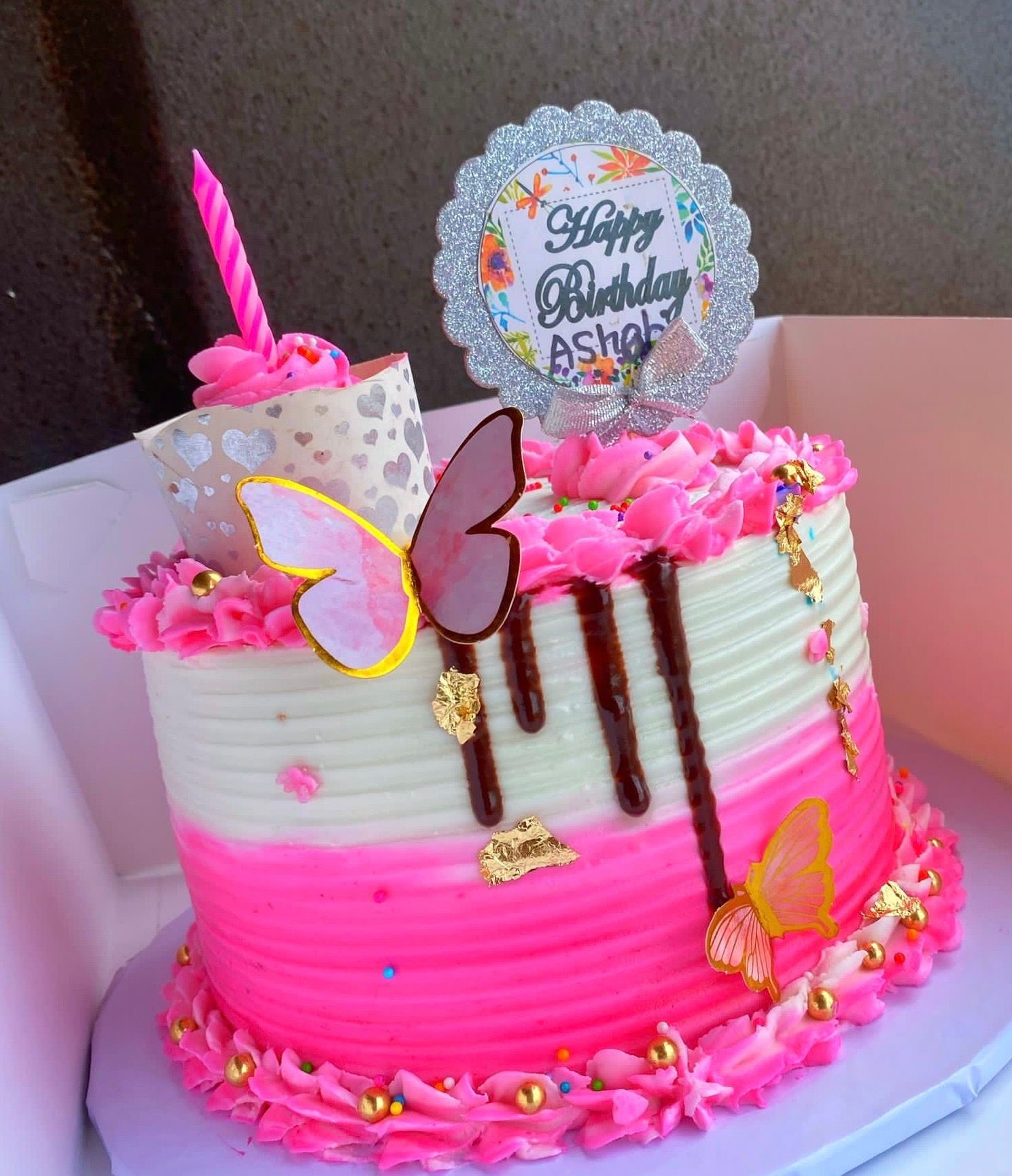 Pink and white birthday cake with gold butterflies, chocolate drips, and a candle.