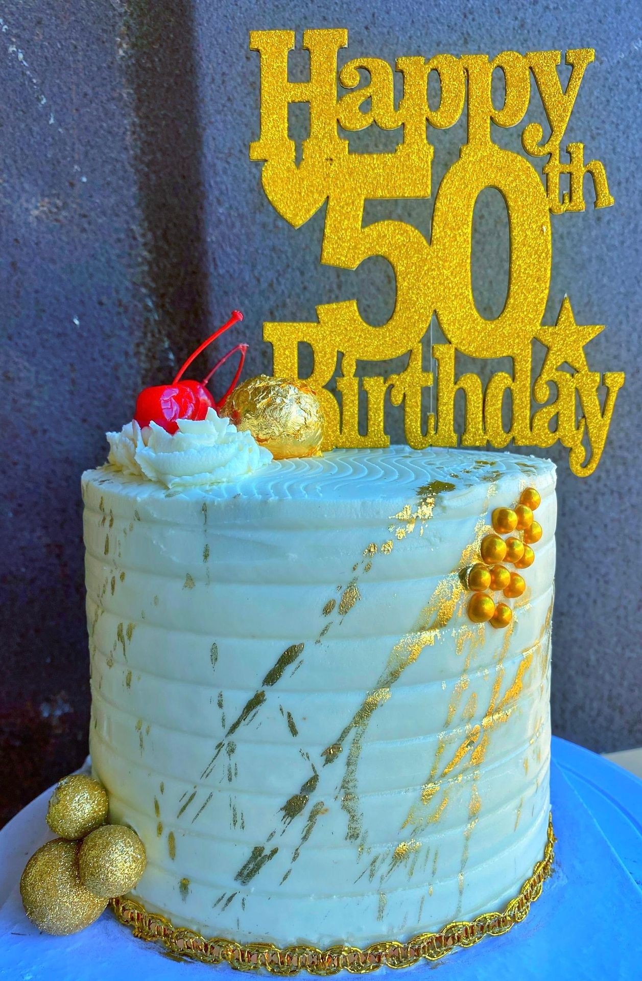 White birthday cake with gold decorations and a 'Happy 50th Birthday' topper.