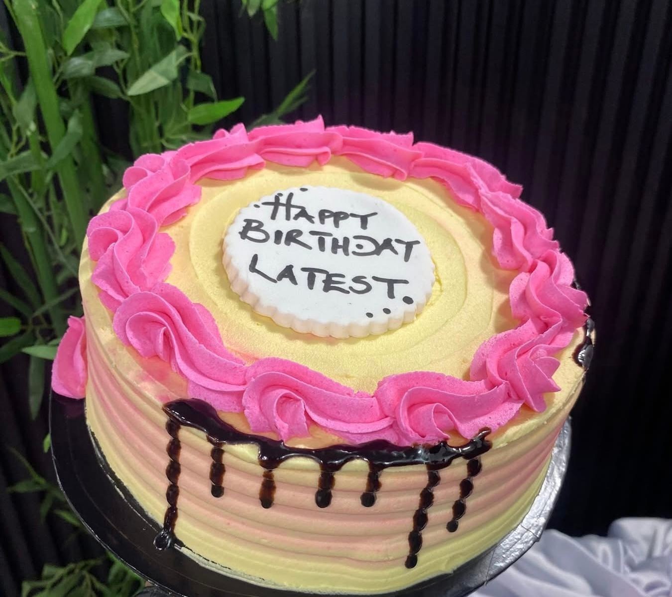 Birthday cake with pink frosting and message 'Happy Birthday Latest' on a white topper.