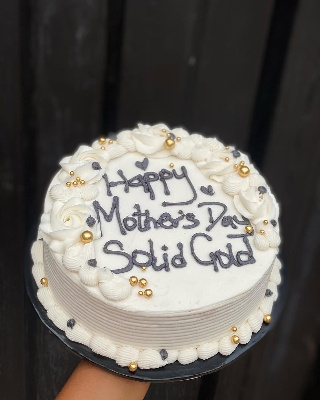 White Mother's Day cake with decorative gold spheres and piped cream details on a dark background.