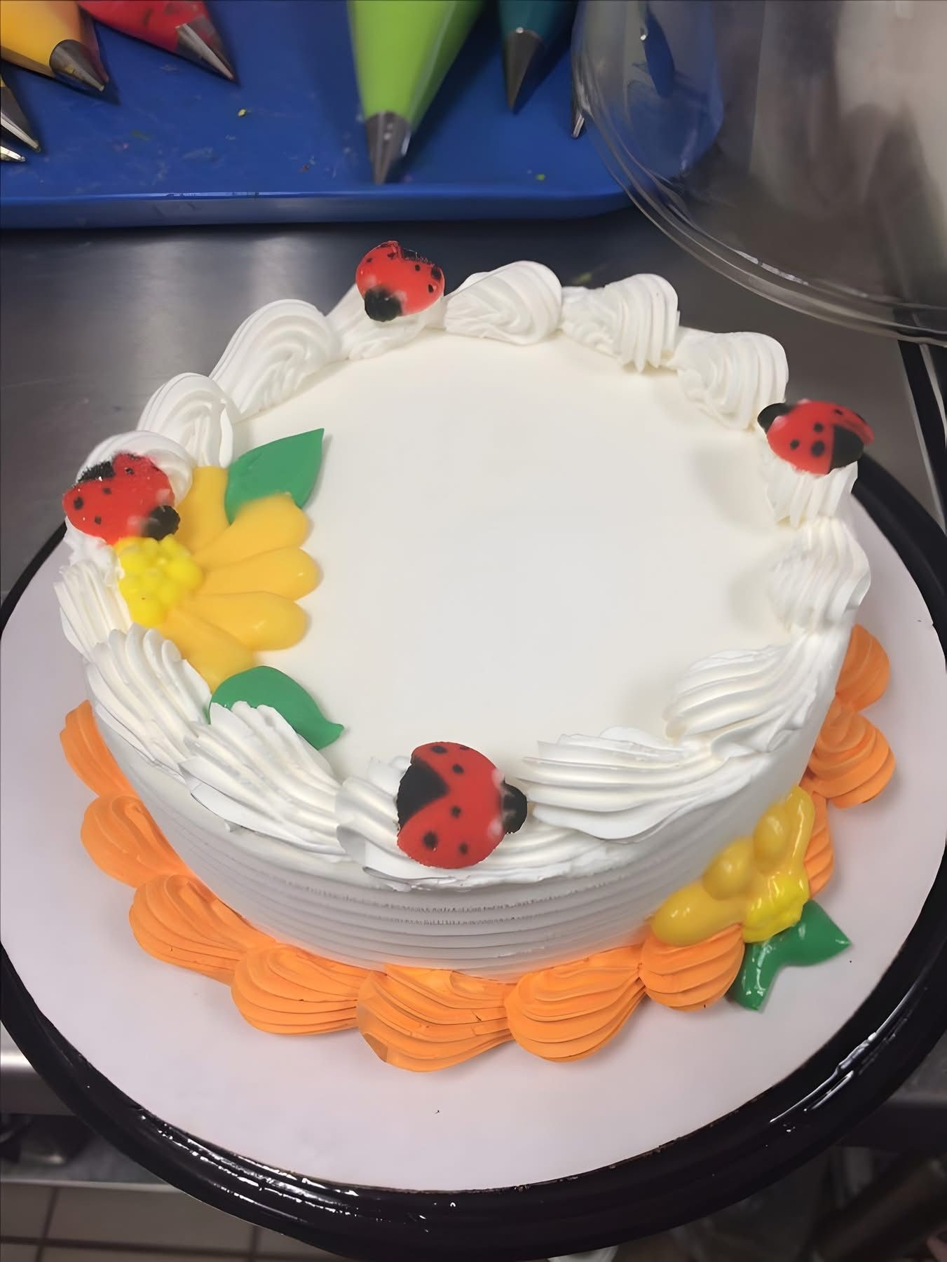 Decorated cake with white frosting, ladybugs, and colorful piping bags in the background.