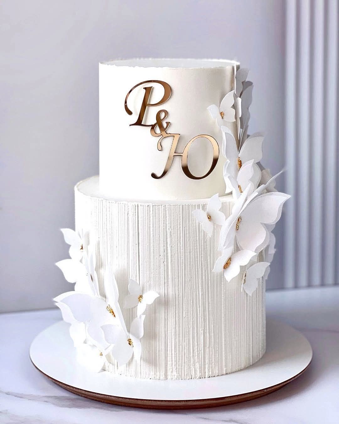 Elegant white two-tier cake with paper flowers and gold letter toppers on a white stand.